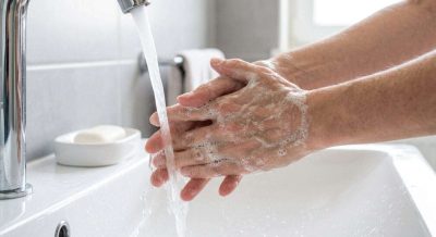 Close-up realistic educational image of hands being properly washed with soap under running water at a clean sink, visible lather covering palms and fingers